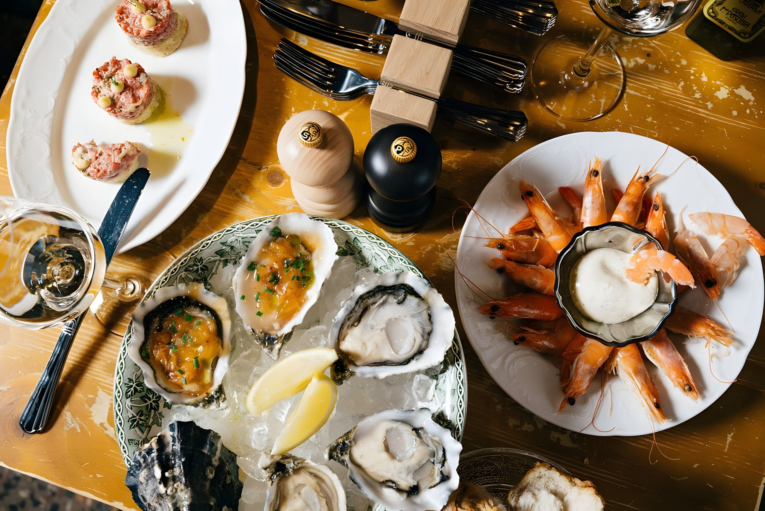Tafel van bovenaf met oesters op ijs, een schaal grijze garnalen met mayonaise en toastjes met tartaar, op een geel geschuurd houten blad